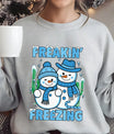 Light gray sweatshirt with “Freakin’ Freezing” DTF transfer featuring two smiling snowmen in blue winter hats and scarves with frosty text, ideal for fun winter apparel.