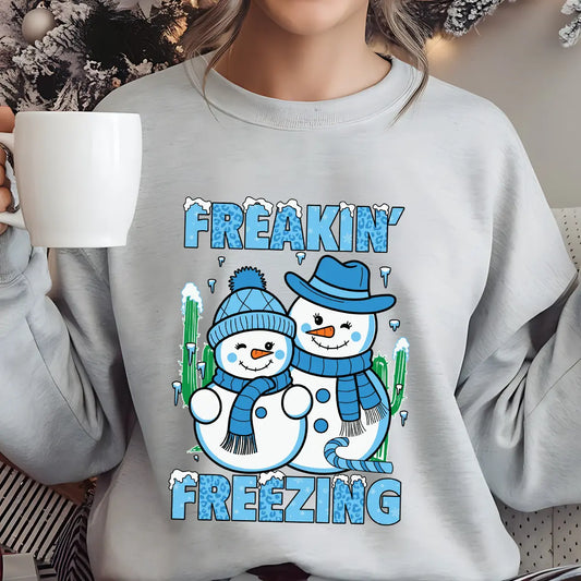 Light gray sweatshirt with “Freakin’ Freezing” DTF transfer featuring two smiling snowmen in blue winter hats and scarves with frosty text, ideal for fun winter apparel.