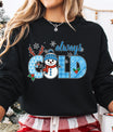 Black sweatshirt with “Always Cold” DTF transfer featuring a smiling snowman, blue plaid letters, snowflakes, holly, and reindeer antlers—perfect for cozy winter wear.