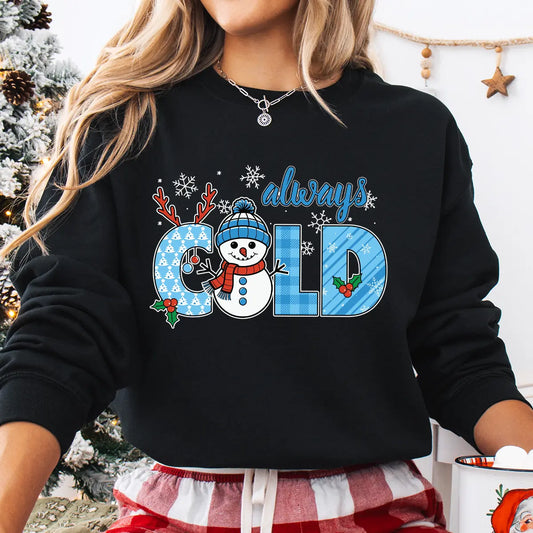 Black sweatshirt with “Always Cold” DTF transfer featuring a smiling snowman, blue plaid letters, snowflakes, holly, and reindeer antlers—perfect for cozy winter wear.