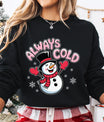 Black sweatshirt with pink “Always Cold” DTF transfer featuring a happy snowman wearing a red scarf and mittens surrounded by snowflakes.