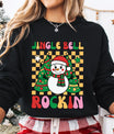 Black sweatshirt with a colorful “Jingle Bell Rockin’” design featuring a smiling snowman and Christmas trees on a yellow checkerboard background.