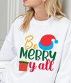 White sweatshirt with colorful “Be Merry Y’all” design featuring a Santa hat, wrapped gift, and festive lettering in red, green, yellow, and blue.