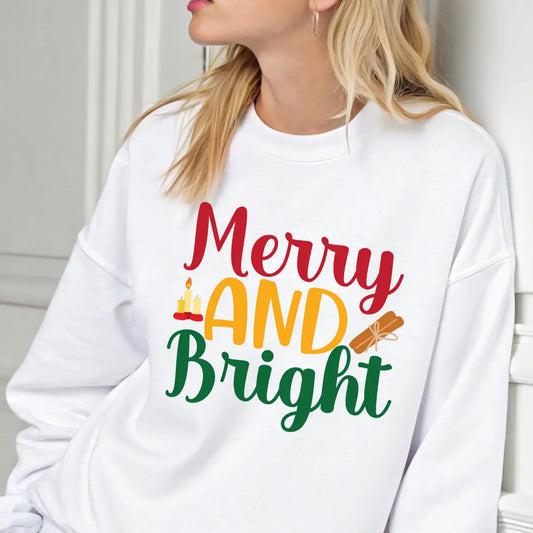 White sweatshirt with “Merry and Bright” text in red, gold, and green featuring candle and cinnamon illustrations – festive holiday apparel.