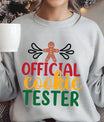 Grey sweatshirt with “Official Cookie Tester” text and gingerbread man design in festive red, gold, and green colors – fun Christmas baking sweatshirt.