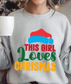 Gray sweatshirt with colorful text “This Girl Loves Christmas” featuring a red Santa hat and festive design elements.