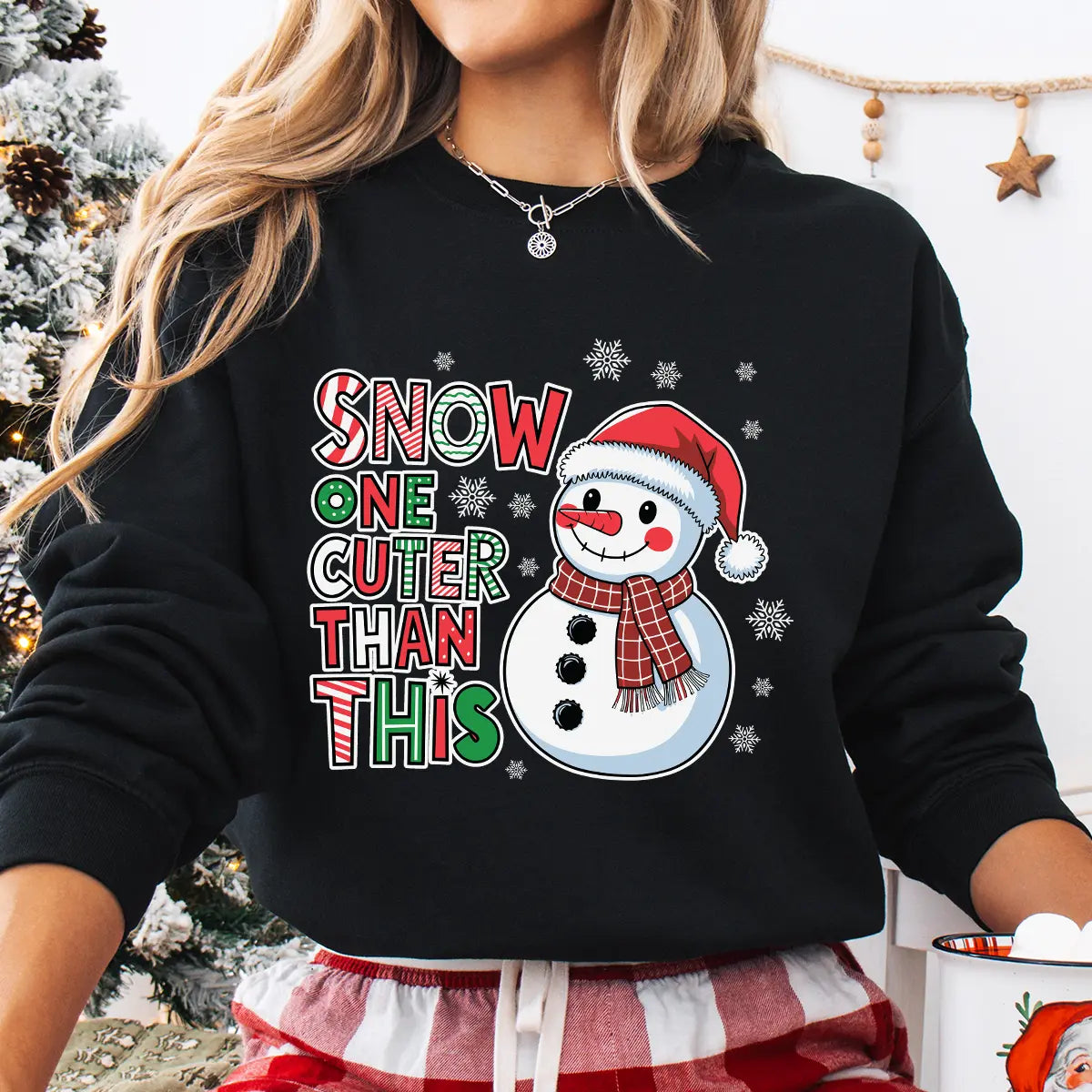 Black sweatshirt featuring a smiling snowman in a Santa hat and plaid scarf with the phrase “Snow One Cuter Than This” in festive red, green, and white lettering.