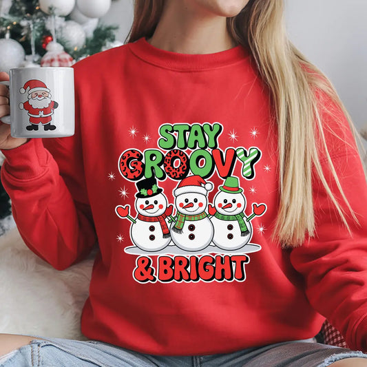 Red sweatshirt featuring three smiling snowmen with scarves and hats and the phrase “Stay Groovy & Bright” in festive red and green lettering, ideal for Christmas apparel.
