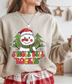 Beige sweatshirt featuring a snowman with a Santa hat and two Christmas trees behind, with colorful text “Jingle Bell Rockin’,” ideal for festive Christmas apparel.