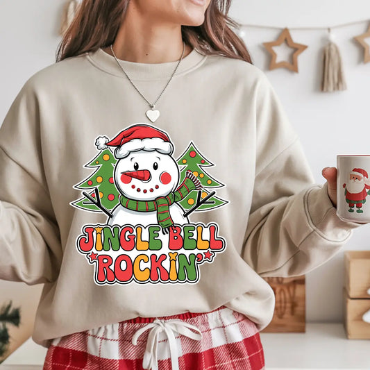 Beige sweatshirt featuring a snowman with a Santa hat and two Christmas trees behind, with colorful text “Jingle Bell Rockin’,” ideal for festive Christmas apparel.