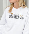 White sweatshirt featuring a black and gold “Believe” DTF design with Santa hat, ornament, and reindeer antlers—minimal and elegant Christmas style.