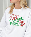 White sweatshirt with “Joy to the World” design in red and green text, decorated with holly leaves, gold stars, and gifts.