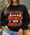 Black sweatshirt with bold orange and white text “This Witch Can Be Bribed with Chocolate” DTF transfer design featuring flying witches and candy graphics.