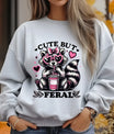 Gray sweatshirt featuring a raccoon with pink heart sunglasses and bow drinking from a cup, with text “Cute But Feral” DTF transfer design.
