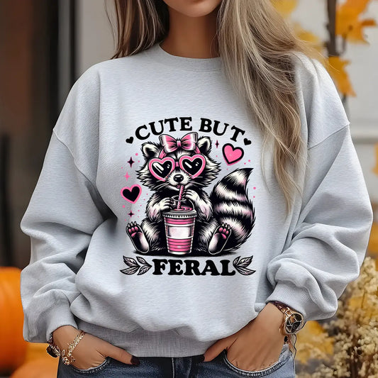 Gray sweatshirt featuring a raccoon with pink heart sunglasses and bow drinking from a cup, with text “Cute But Feral” DTF transfer design.