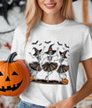 White T-shirt with three dancing skeleton witches wearing hats and skirts, surrounded by bats and pumpkins, Halloween DTF transfer design.