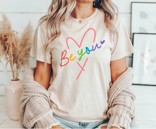 Be You Rainbow Heart DTF Transfer design on a cream T-shirt worn by a woman.