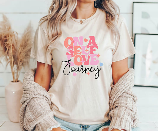 On a Self Love Journey DTF Transfer design with retro-style pastel lettering and heart accents on a cream T-shirt worn by a woman.