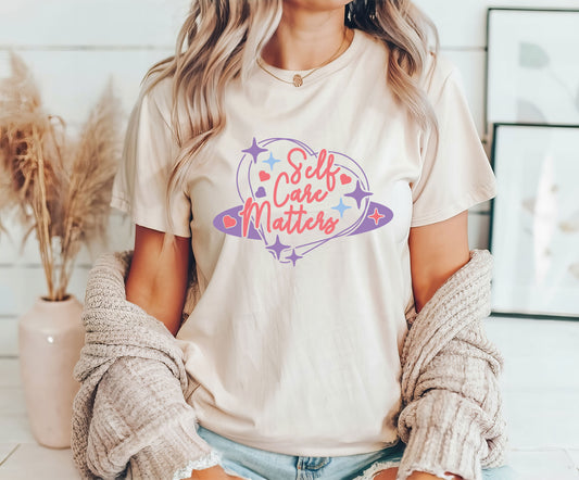 Self Care Matters DTF Transfer design in pastel tones with stars and a heart shape on a cream T-shirt worn by a woman.