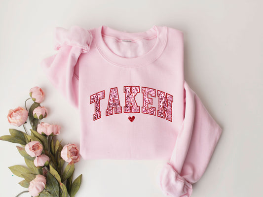 TAKEN Valentine's Day ready-to-press DTF Transfer design with glittery red letters and a heart on a pink sweatshirt.